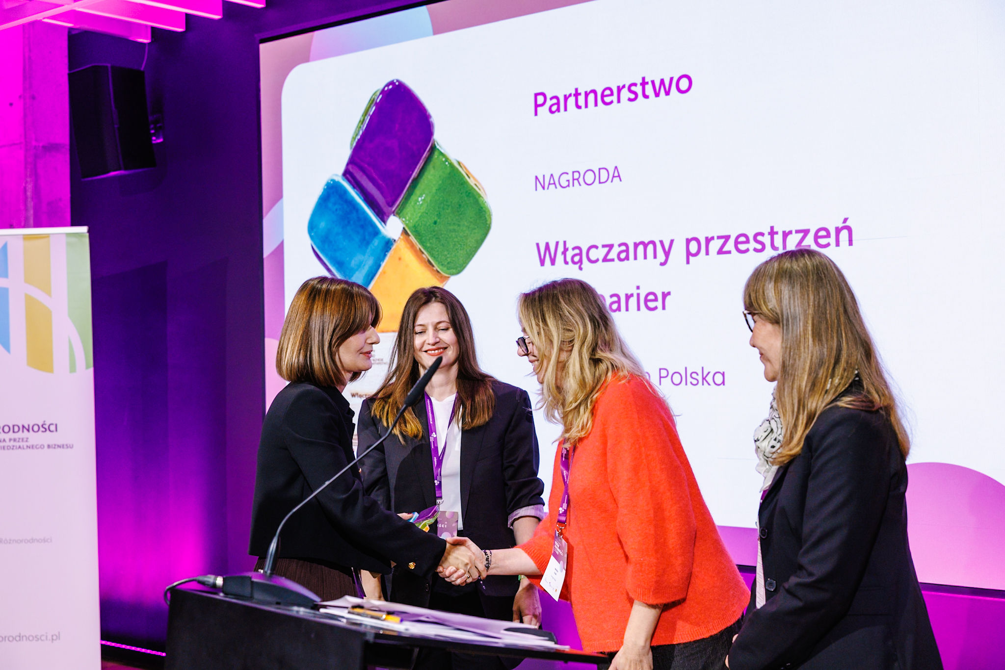 Cztery kobiety na scenie podczas gali Karty Różnorodności. Dwie osoby podają sobie rękę przy mikrofonie. W tle ekran z napisem „Partnerstwo. Nagroda. Włączamy przestrzeń bez barier. Leroy Merlin Polska.”